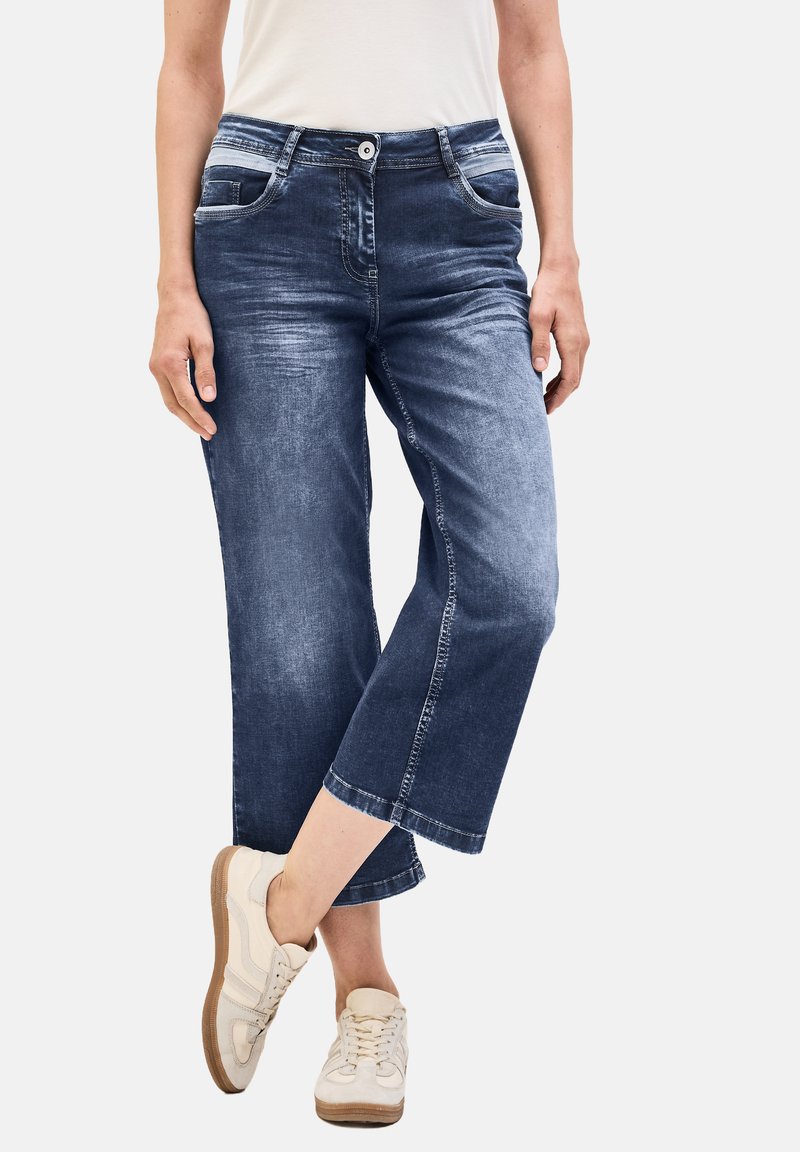 Culottes en denim bleu foncé à finition délavée, dotées de poches latérales, d'une fermeture à bouton et d'une coupe droite. Portés avec des baskets claires.