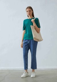 Kortermet topp i blågrønn, blå jeans med rette ben og hvite joggesko. Beige handlenett hengt over skulderen. Enkel design, ensfargede plagg.