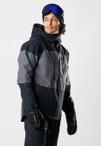 Homme portant une veste de ski noire et grise, des gants noirs, un pantalon noir et un masque de ski bleu sur un bonnet noir, regardant vers sa droite.