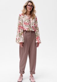 Bloemenblouse met lange klokmouwen, roze en groen op een crèmekleurige achtergrond; hooggetailleerde beige broek; lichtroze sneakers.