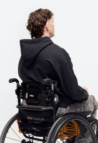 Personne aux cheveux bouclés portant un sweat à capuche noir assise dans un fauteuil roulant manuel avec des rayons de roue orange, vue de dos sur fond blanc.