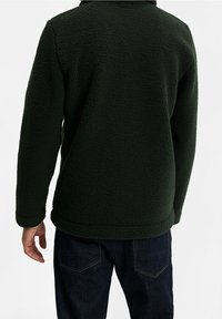 Tmavě zelená fleecová bunda s texturovaným, jemným povrchem, límcem a dlouhými rukávy. Nositá s námořnickými modrými džíny.