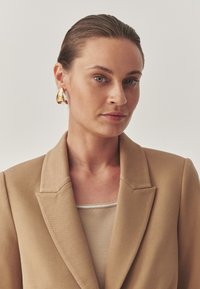 Beige blejzr s klopovými límci přes světle béžový top. Zlaté kruhové náušnice přidávají výrazný detail. Hladká, strukturovaná textura látky.