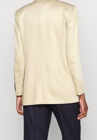 Blazer beige con una textura suave, diseño ajustado y corte recto; presenta un sutil acolchado en los hombros y acentos de botones negros en los puños.