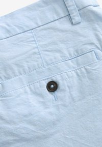 Shorts en tissu bleu clair à texture lisse, avec une poche arrière munie d'un rabat à bouton marron et des détails cousus.