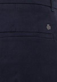 Tumši zili bikses, izgatavotas no kokvilnas maisījuma; tās ir ar aizmugures kabatu un mazu apaļu zīmola logotipu labajā pusē. Strukturēta auduma.