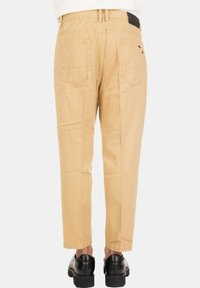 Pantaloni beige dritti con tasche posteriori indossati con scarpe in pelle nere da una persona in piedi davanti a uno sfondo bianco.