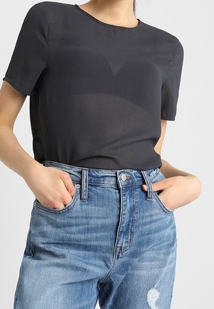 Femme portant un haut noir à manches courtes rentré dans un jean bleu taille haute, mains dans les poches avant, sur un fond uni.