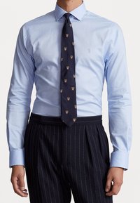 Světle modrá košile s límečkem na knoflíky, doplněná tmavě modrou kravatou se vzorem štítu a černé kalhoty s tenkými proužky.