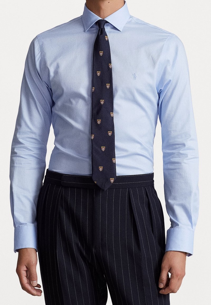 Světle modrá košile s límečkem na knoflíky, doplněná tmavě modrou kravatou se vzorem štítu a černé kalhoty s tenkými proužky.