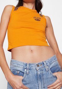 Orange croppad linne med bruna axelaccenter, med "BDG Jeans"-logotyp i mörk skrift. Bärs med ljusblå hög midja jeans.