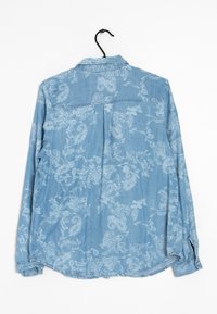 Camicia in denim azzurro chiaro con un intricato motivo paisley bianco, polsini a bottoni, colletto e dettaglio con piega sul retro. Appesa a un gancio nero.