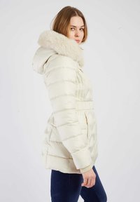 Witte donzenjas met een capuchon gevoerd met bont, geribbelde manchetten en een taille met trekkoord. Heeft gouden ritssluitingen op de zakken en een doorgestikt patroon.