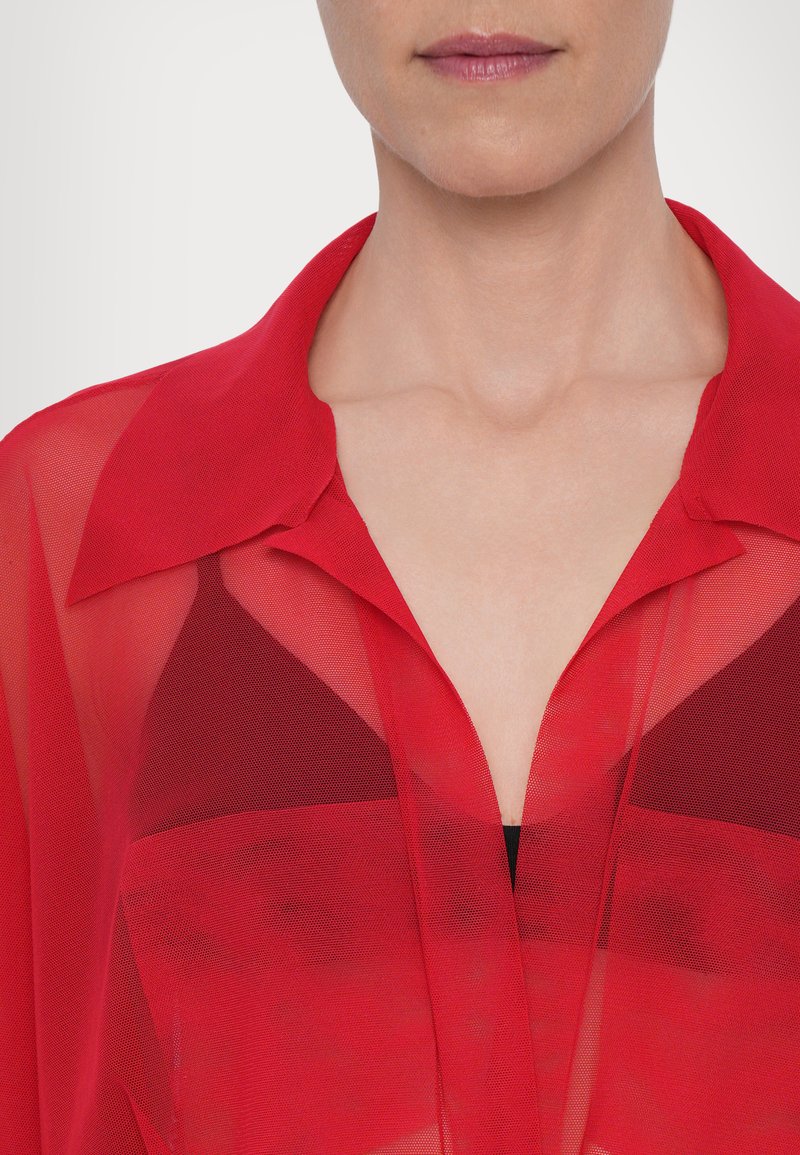 Camicia rossa traslucida con colletto, caratterizzata da una trama semi-trasparente. Il design include un scollo a V profondo, sovrapposto a una maglietta nera.