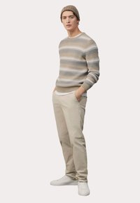 Gestreifter Strickpullover in Beige- und Grautönen, kombiniert mit hellbeigen Hosen und weißen Sneakern. Das Model posiert mit den Händen in den Taschen.