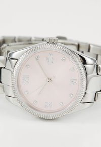 Silverfärgad klocka i rostfritt stål med en rosa urtavla, prydd med sex diamantlika markörer, räfflad bezel och Lacoste-logotyp vid klockan 12.