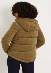 Chaqueta acolchada de pana marrón con capucha, con una textura acolchada y un dobladillo ligeramente curvado, llevada sobre vaqueros oscuros.