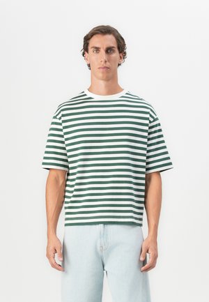 Young man with short brown hair wearing a green and white striped short-sleeve T-shirt and light blue jeans, standing against a white background.