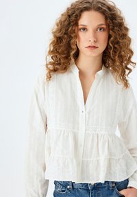 Witte blouse met knopen en horizontale plooien en kantafwerking; heeft lange mouwen en een flared, gerimpelde zoom. Draagt met blauwe jeans.