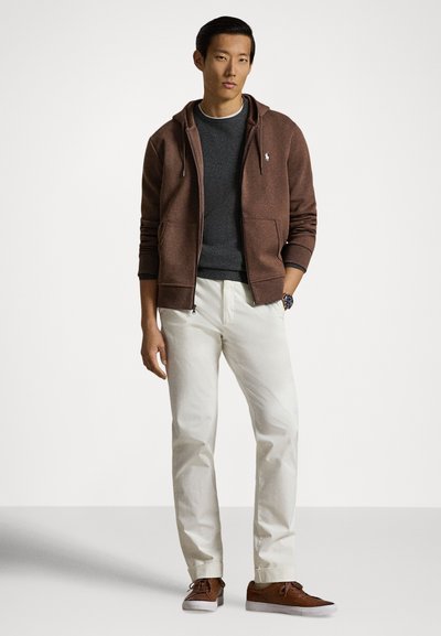 Brown zip-up hoodie over a dark gray sweater, paired with white pants and brown sneakers. Features front pockets and a logo on the hoodie.