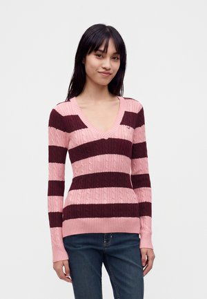 Vrouw met een lang mouw roze en donkerrode gestreepte V-hals trui en blauwe spijkerbroek, staand tegen een eenvoudige lichte achtergrond.