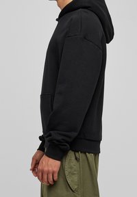 Sudadera negra con capucha y un bolsillo frontal, con puños y dobladillo acanalados, fabricada con un material suave y texturizado. Combinada con pantalones verde oliva.