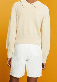Personne portant un pull crème à manches longues avec un large col et un short blanc taille haute sur fond orange.