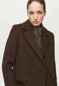 Cappotto marrone doppiopetto con collo a revers, dotato di due tasche frontali e bottoni neri, indossato sopra un maglione con cerniera testurizzato.