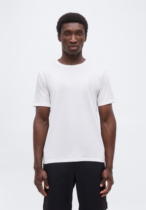 Ung mand stående vendt fremad iført en ensfarvet hvid kortærmet t-shirt og sorte bukser mod en ensfarvet lys baggrund.