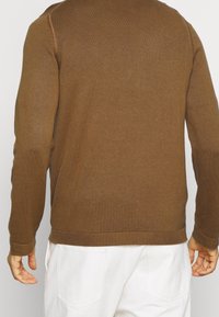 Brun stickad tröja med ribbad textur, långa ärmar och en åtsittande fåll. Bärs med vita byxor, visar bakifrån.