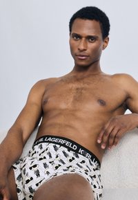 Baltos bokserinės kelnaitės su juodu juosmens diržu, ant kurio parašyta „LAGERFELD KORL“. Ant audinio žaismingai išdėstyti animacinių personažų su akiniais nuo saulės raštai.