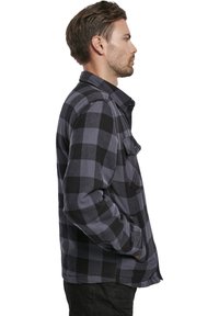 Camisa de franela a cuadros en negro y gris con cuello, bolsillo frontal y diseño de botones. La tela parece suave y texturizada.
