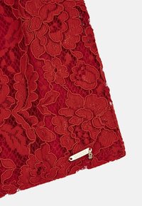 Tissu en dentelle rouge avec motifs floraux, broderies intriquées, et une petite étiquette métallique avec l'inscription "GUESS" sur le bord inférieur.