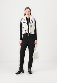 Krämfärgad, texturerad väst med blå blommönster, bärs över en svart skjorta och svarta jeans. Modellen håller en vit handväska och har på sig svarta klackstövlar.