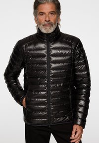 Svart puffjacka med hög krage, med quiltade sektioner, fram zip och logotypdetalj på vänster bröst. Mjuk, glänsande textur.