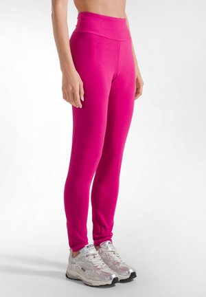 Vrouw die felroze hoge taille leggings en zilveren en witte sneakers draagt, staand op een witte achtergrond.