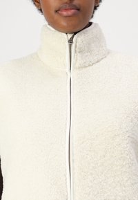 Vit fleecejacka med hög krage, hel dragkedja och texturerad yta. Mjuk material och kontrasterande dragkedjefärg. Inga synliga mönster.