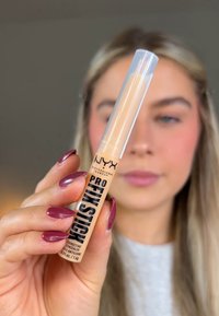 NYX Pro Finishing Stick, beige, contenitore di plastica, cappuccio trasparente, testo nero in grassetto, tenuto in mano con unghie corte, finitura liscia e opaca.