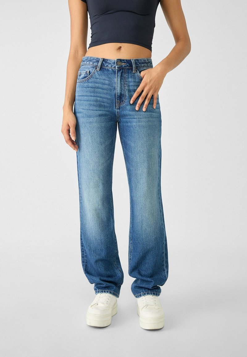 Femme portant un jean denim bleu taille haute avec des détails délavés, des baskets blanches et un top court noir, debout devant un fond uni.
