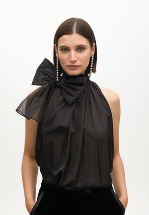 Black halter top with a large sheer bow at the neckline, featuring a textured accent, paired with long pearl earrings. Smooth fabric.