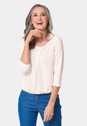 Un haut crème clair à manches trois quarts, avec un col rond et un léger froncé à l'avant. Assorti à un jean en denim bleu.