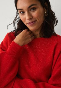 Rød strikket sweater med rund halsudskæring, tekstureret mønster og diskrete dekorationer. Har ribbede manchetter og en afslappet pasform.