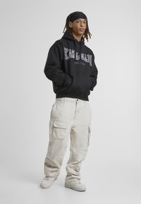 Felpa nera con grafiche bianche, abbinata a pantaloni cargo beige dalle linee morbide e scarpe da ginnastica bianche. Tessuto liscio, design rilassato.