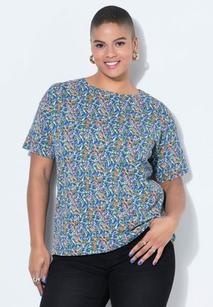 Lächende Person mit kurzem Haar, die goldene Creolen, ein mehrfarbiges, kurzärmeliges Oberteil mit Blumenmuster, schwarze Hose und blaue Nagellack trägt.