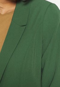 Green blazer with a smooth texture and structured lapels, paired with a brown top. Features a relaxed fit and minimal stitching details.