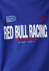 Μπλε ύφασμα με λευκό και κόκκινο κείμενο που γράφει «RED BULL RACING Ανταγωνιζόμενοι από το 2006» και μικρότερα λογότυπα «Racing Gear» και «Team Racing».