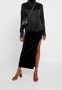Schwarze Satin-Bluse mit langen Ärmeln und hohem Kragen, kombiniert mit einem schwarzen Samtrock mit einem Oberschenkelspalt. Beigefarbene, strappy High Heels runden das Outfit ab.