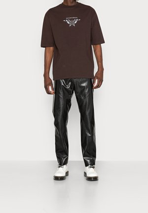 Brun oversized t-shirt med vit grafisk design, kombinerad med svarta glansiga byxor i syntetläder och vita skor med svarta sulor.