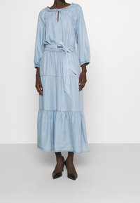 Ljusblå denimklänning med långa ärmar, volangkrage, nyckelhålöppning, åtdragen midja och tierad nederkant. Svarta högklackade skor.