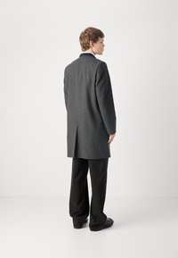 Grauer Wollmantel mit geradem Schnitt, mit einem Schlitz hinten und schwarzen Revers, kombiniert mit schwarzen Hosen und Schuhen.